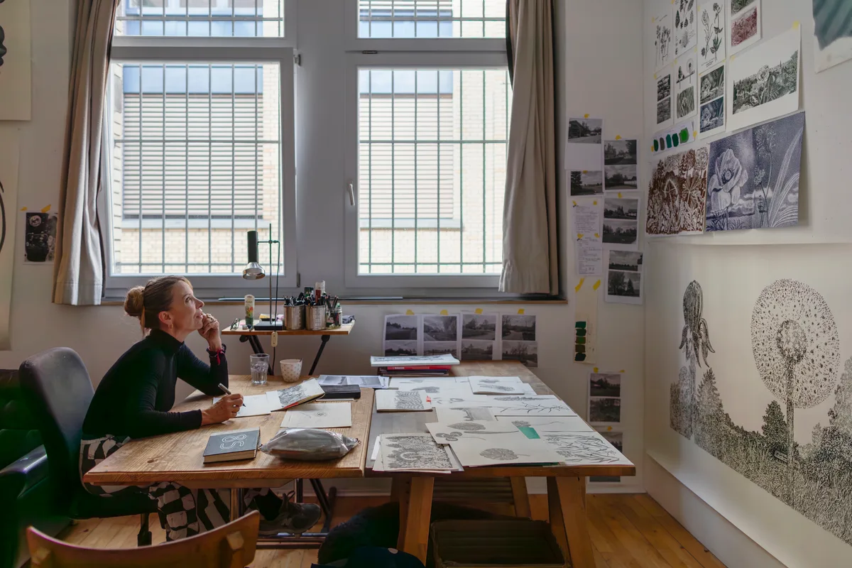 Eine Künstlerin sitzt an einem Tisch in ihrem Atelier und schaut an die Wand, wo mehrere Skizzen hängen.